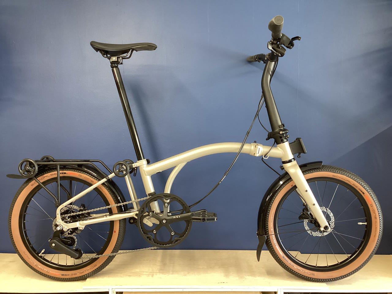 A Brompton G Line bicycle with a roller frame, in Traildust White color, folded and placed upright against a blue wall on a white floor.